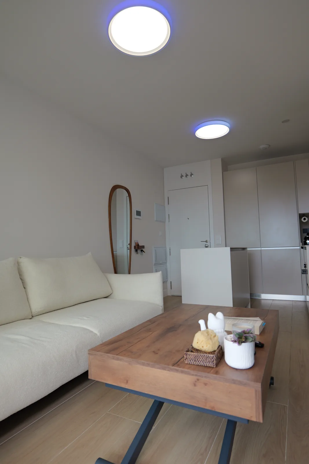 Salón con sofá claro, mesa de madera y acceso a la cocina con plafón de luz en el techo
