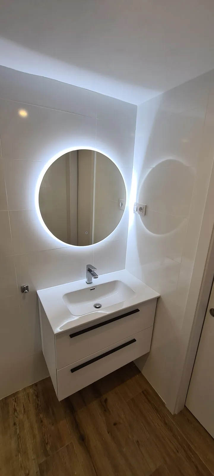 Lavabo blanco suspendido con espejo redondo retroiluminado y suelo imitación madera