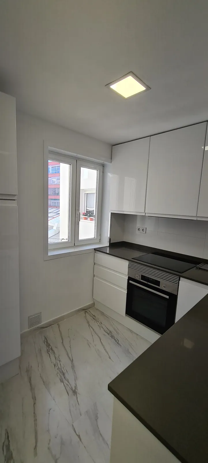 Cocina blanca brillo con encimera gris oscura y ventana al fondo