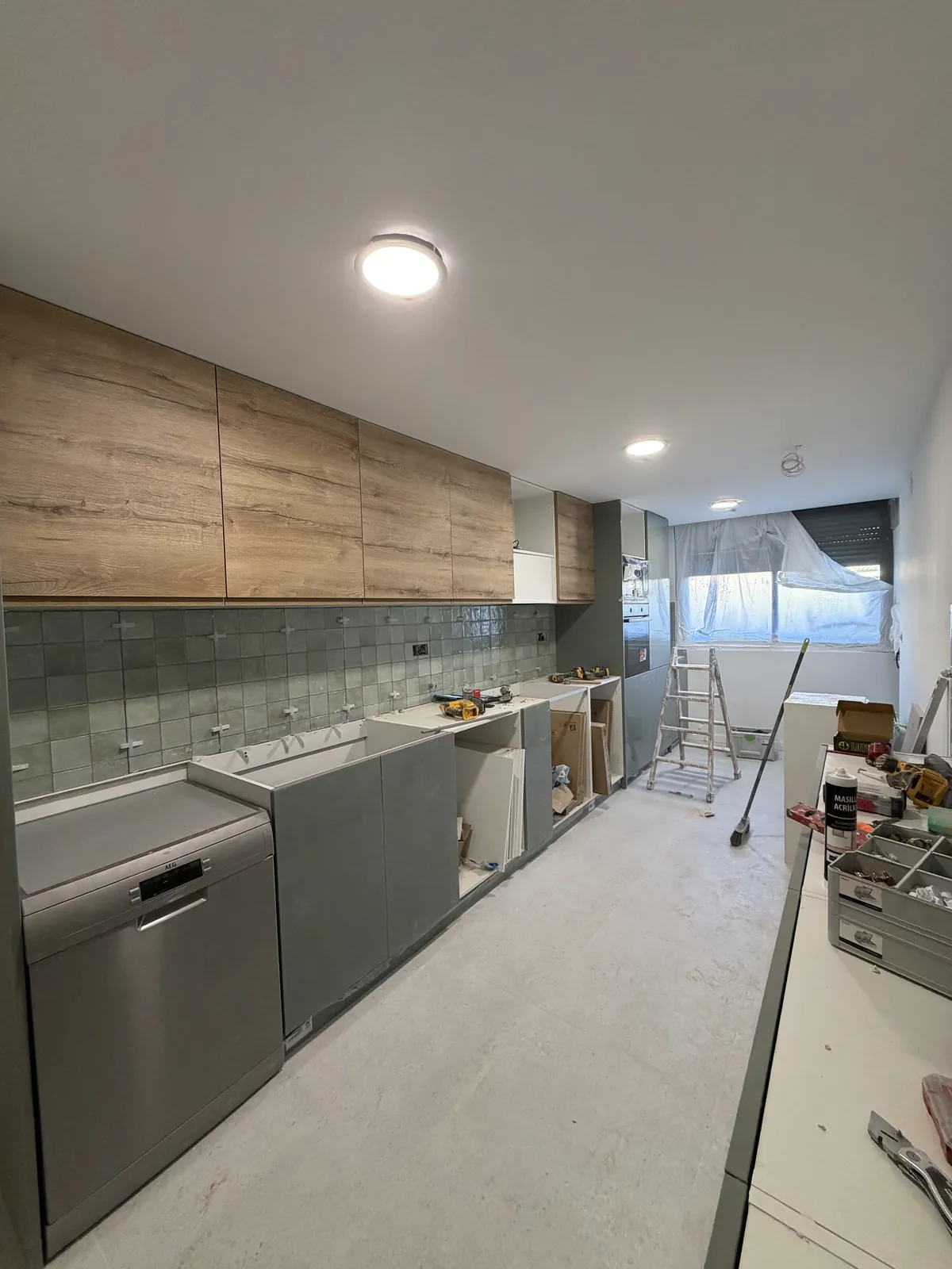 Cocina en montaje con muebles altos en madera, bajos verde grisáceo y frontal de azulejo verde