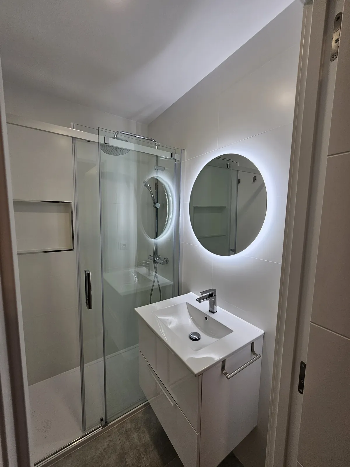 Baño con mampara corredera, lavabo blanco y espejo redondo retroiluminado