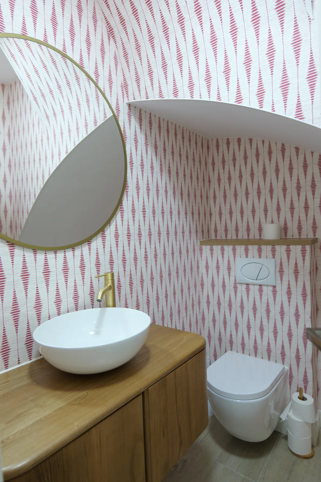 Aseo con papel pintado en rosa, mueble de madera con lavabo sobre encimera, grifo dorado e inodoro suspendido