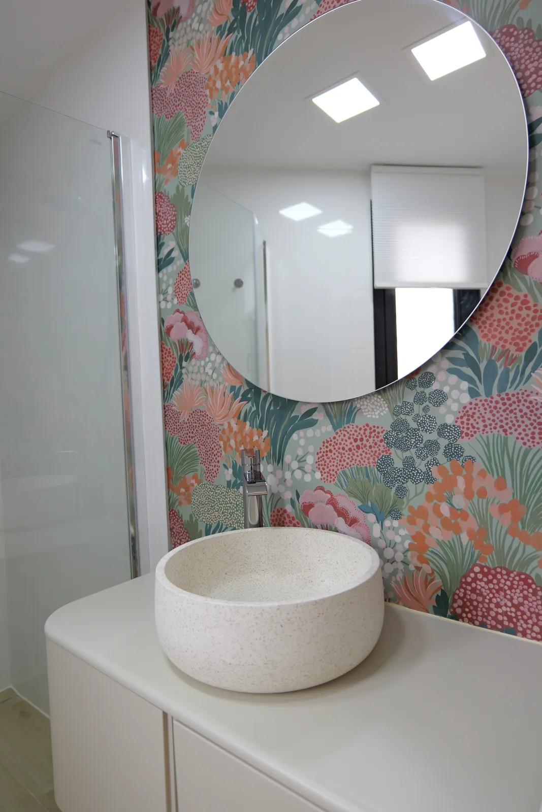 Baño con papel pintado floral, lavabo de piedra sobre mueble lacado y espejo redondo