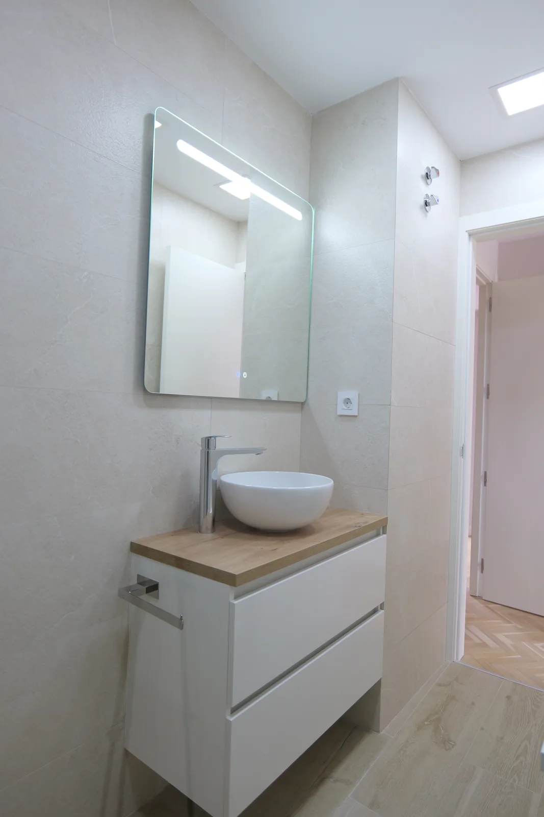 Baño en tonos claros con mueble blanco y encimera de madera, lavabo de sobre encimera y espejo con luz LED