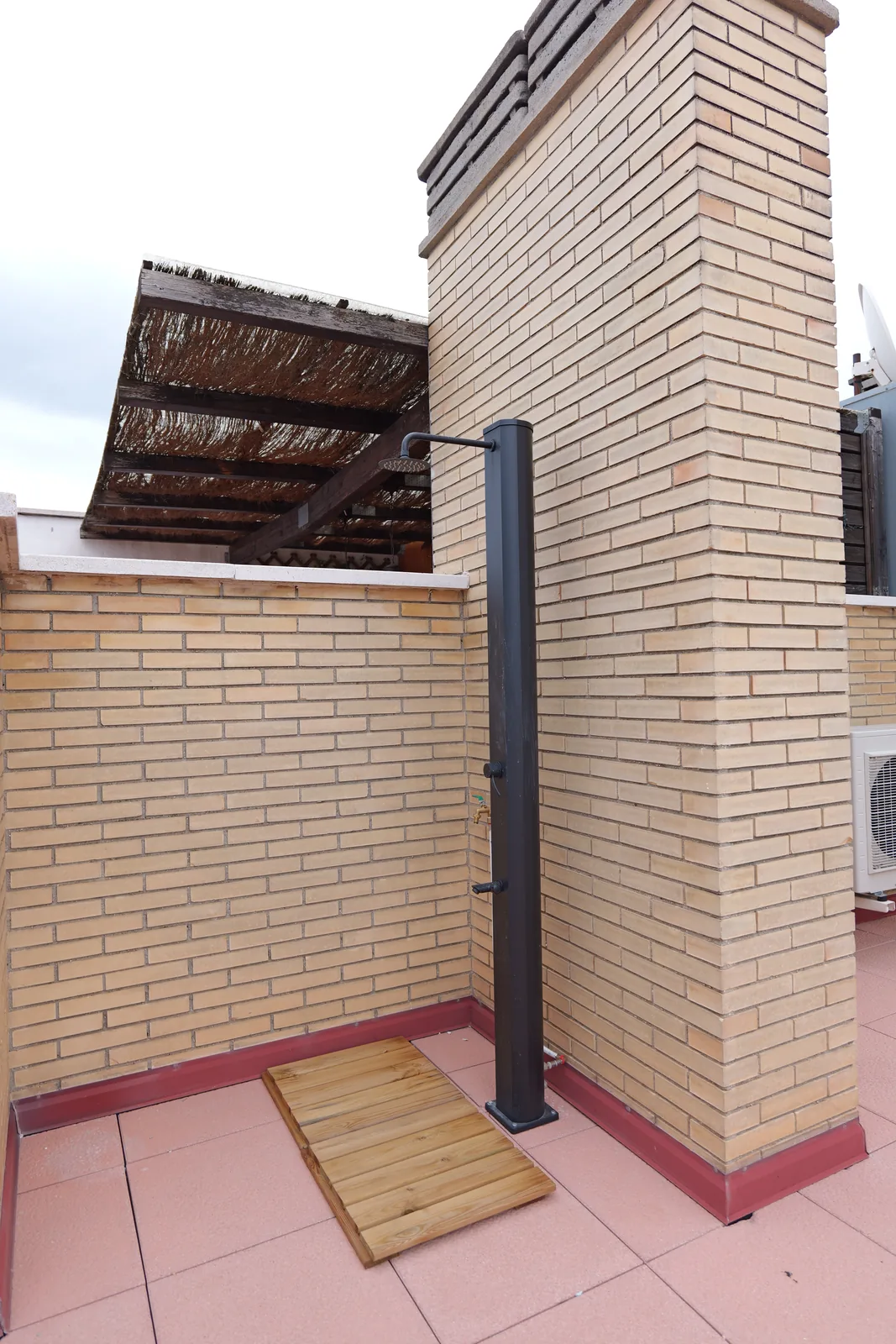 Ducha exterior de columna en la terraza junto a muro de ladrillo visto y tarima de madera