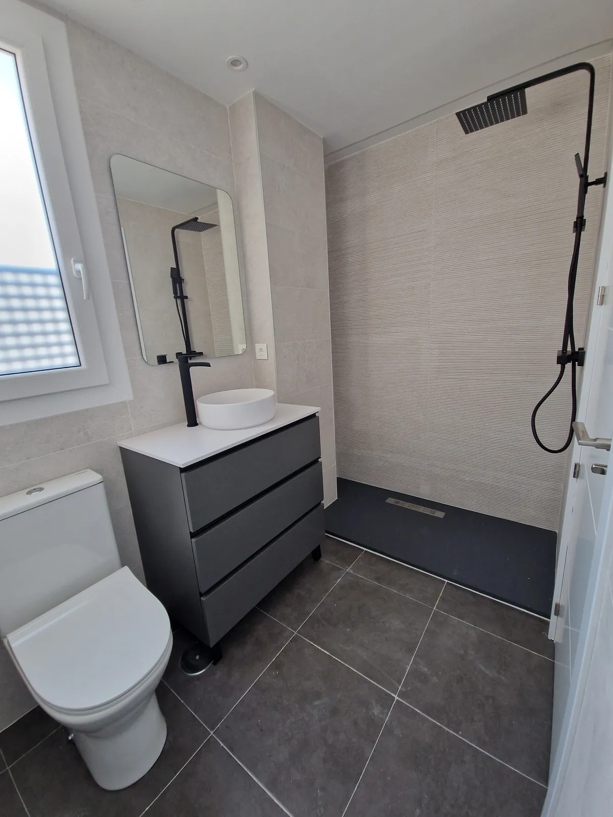 Baño con columna de ducha negra, mueble de lavabo gris y revestimiento porcelánico claro