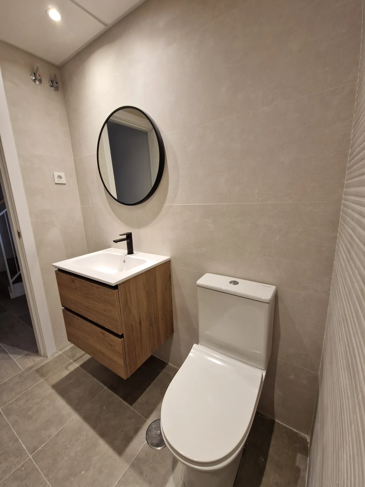 Baño con mueble suspendido de madera, espejo redondo de marco negro e inodoro blanco