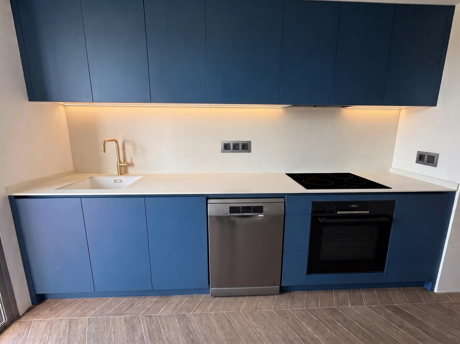Cocina reformada con muebles azul mate, encimera blanca, fregadero bajo encimera, lavavajillas y horno integrados