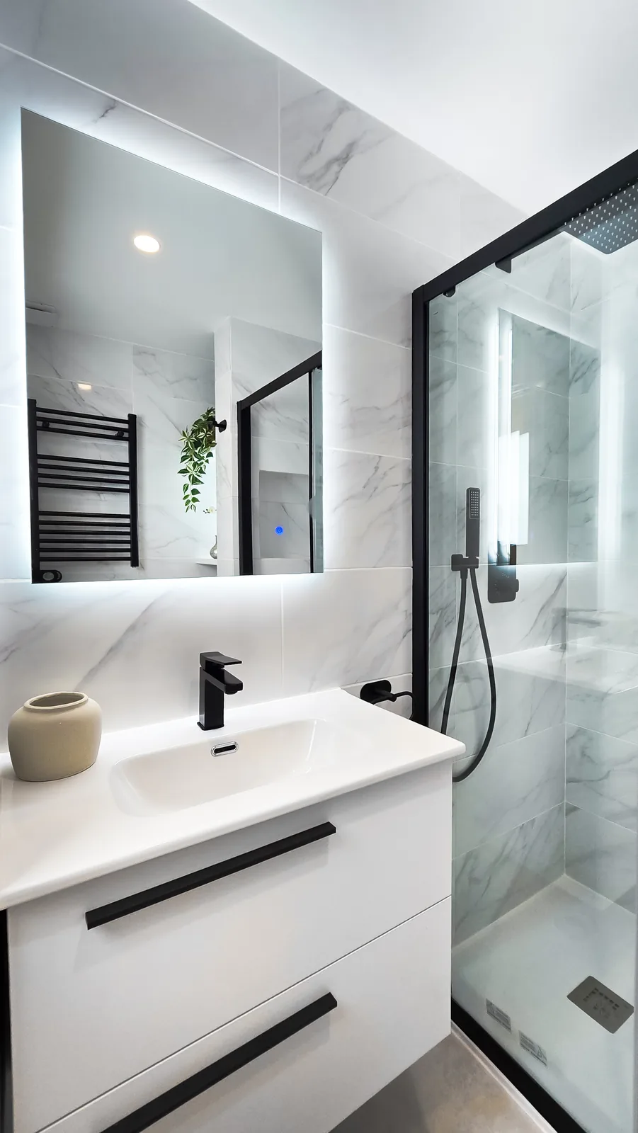 Baño reformado con mueble de lavabo blanco, grifería negra, espejo retroiluminado y mampara negra