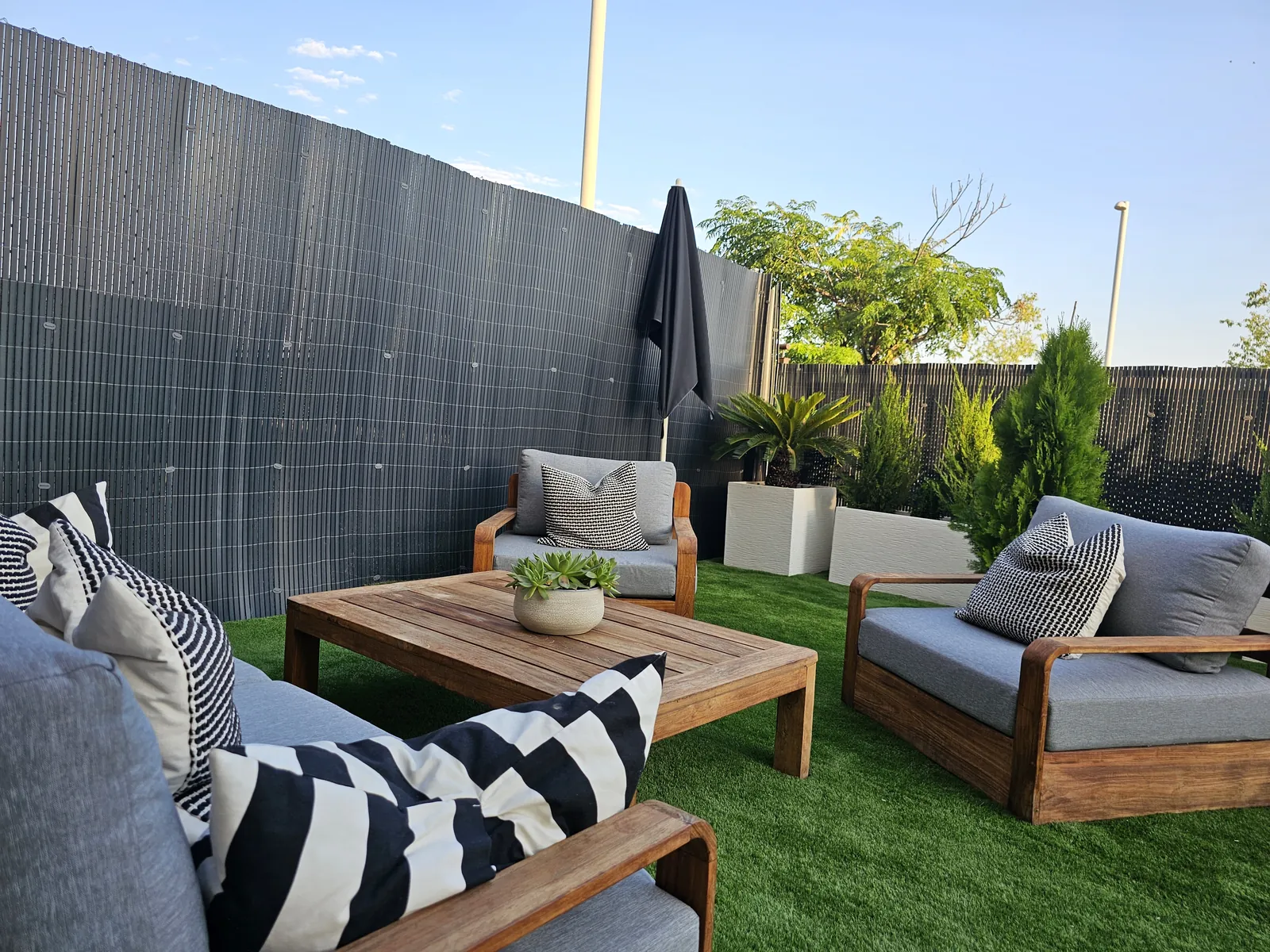 Patio con césped artificial, valla gris oscuro y zona de estar con sofás de madera