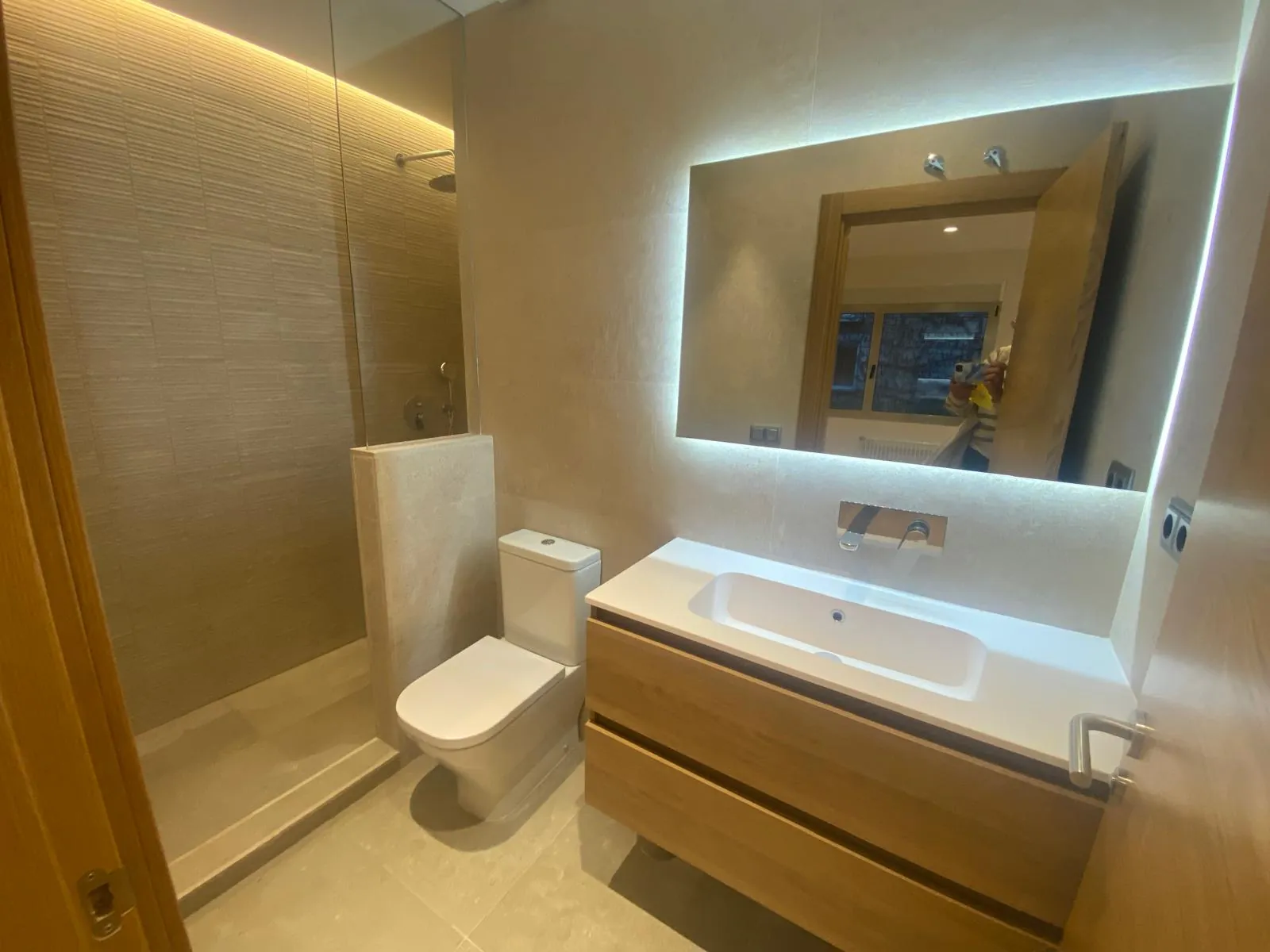 Baño con mueble de madera suspendido, espejo retroiluminado y ducha de obra