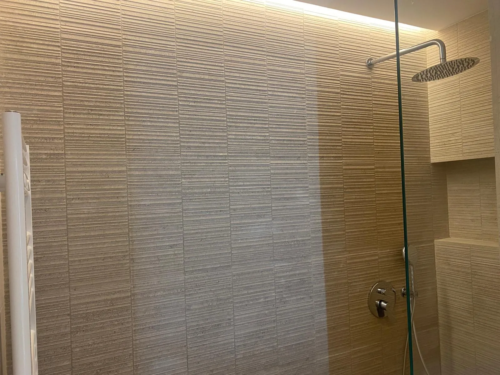 Ducha con revestimiento beige texturizado, rociador de techo y luz indirecta